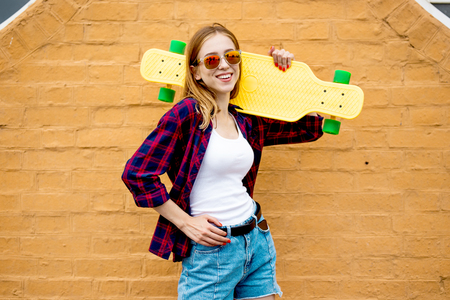 A pretty blond smiling girl wearing sunglasses, checkered shirt and denim shorts is standing in front of the brick wall and holding a yellow longboard on her shoulder. Cool style.の写真素材