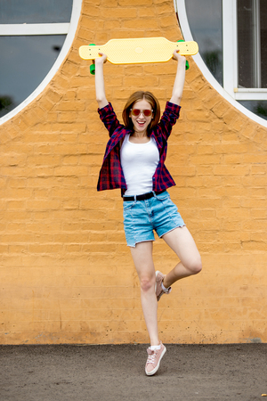 A pretty blond smiling girl wearing sunglasses, checkered shirt and denim shorts is standing on one leg in front of the brick wall and holding a yellow longboard above her head. Cool style.の写真素材
