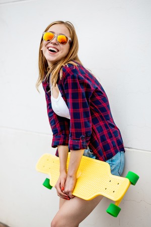 A pretty smiling blond girl wearing sunglasses, checkered shirt and denim shorts is standing in front of the gray wall and holding a yellow longboard in front of her. Cool style.の写真素材