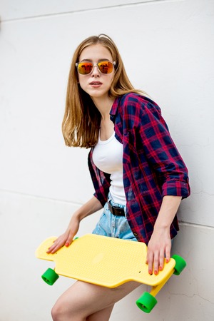 A pretty blond girl wearing sunglasses, checkered shirt and denim shorts is standing in front of the gray wall and holding a yellow longboard in front of her. Cool style.の写真素材