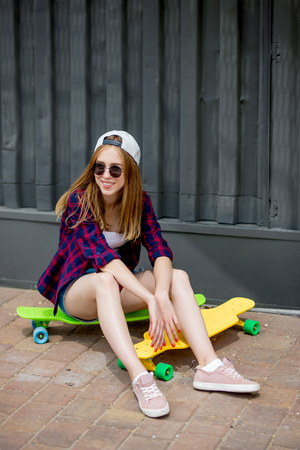 A pretty smiling blond girl wearing sunglasses, checkered shirt and denim shorts is sitting on the bright logboards in front of the black wall. Cool style.の写真素材