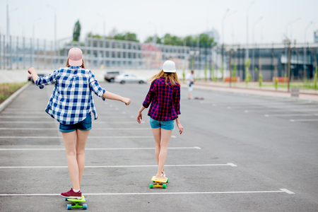 Two blond girls wearing checkered shirts, caps and denim shorts are longboarding on the empty car park. Sport and cool style.の写真素材