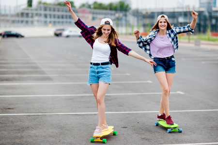 Two pretty smiling blond girls wearing checkered shirts, caps and denim shorts are longboarding on the empty car park with their arms stretched. Sport and cool style.の写真素材