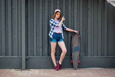A pretty blond girl wearing checkered shirt and denim shorts is standing in front of the black wall with a black longboard next to her. Cool style.の写真素材