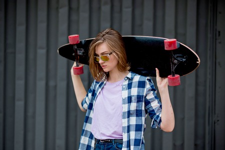 A pretty smiling blond girl wearing checkered shirt and denim shorts is standing in front of the black wall holding a black longboard on her shoulders. Cool style.の写真素材