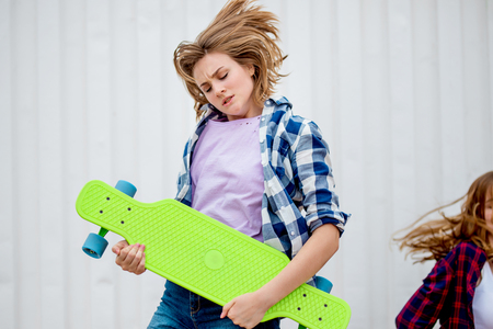 A pretty blond girl wearing checkered shirt and denim shorts is standing in front of the gray wall holding a grin longboard in front of her. Flying hair. Cool style.の写真素材