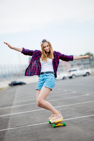 A beautiful blond girl wearing checkered shirt, cap and denim shorts is longboarding while stretching out her hands. Sport and cool style.の写真素材