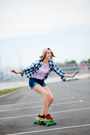 A beautiful smiling blond girl wearing checkered shirt, cap and denim shorts is longboarding while stretching out her hands. Sport and cool style.の写真素材