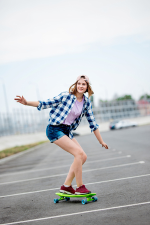 A beautiful smiling blond girl wearing checkered shirt, cap and denim shorts is longboarding while stretching out her hands. Sport and cool style.の写真素材