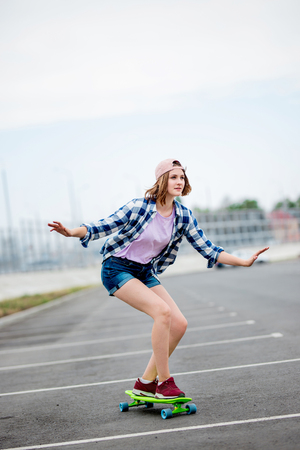 A beautiful smiling blond girl wearing checkered shirt, cap and denim shorts is longboarding while stretching out her hands. Sport and cool style.の写真素材