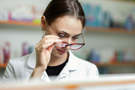 A young focussed brown-haired girl with glasses, wearing a lab coat, stands in a pharmacy by holding her glasses and looks at something.の写真素材