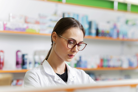 A good looking slim girl with dark hair and glasses,wearing a white coat, gently places goods on the shelf in the pharmacy.の写真素材