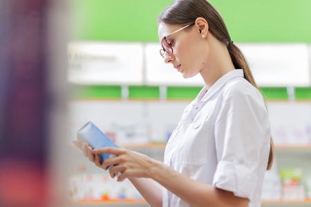 A young thin brown-haired girl with glasses, dressed in a medical overall, reads something on the pills pack next to the shelves in the pharmacy.の写真素材