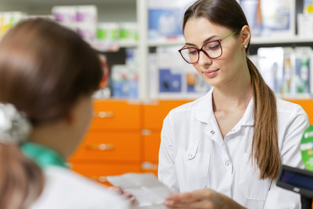 A friendly slim lady with  dark hair and glasses,wearing a lab coat,is talking with a visitor and reads a prescription in a new stylish pharmacy.の写真素材