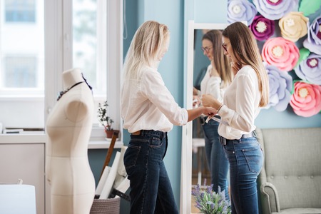 Two smart-looking pretty women wearing white shirts are standing in front of the mirror and measuring each other. Fashion, tailors workshop.の写真素材