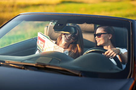 Two dark-haired young women in sunglasses  are sitting in a black cabriolet on a sunny day. One of them keeps map in her hands.の写真素材
