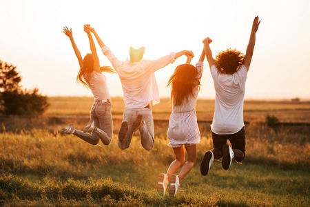 Two young guys and two girls are holding their hand and jumping in the field on a summer day. Back view.の写真素材