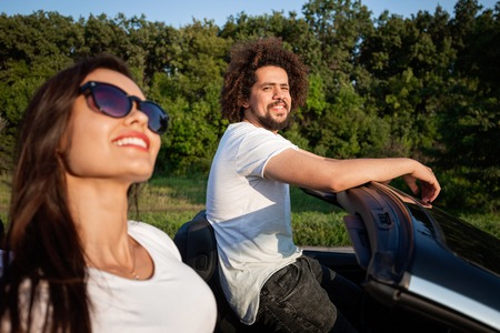 Gorgeous young dark-haired woman in sunglasses with curly young man are sitting in a black cabriolet...