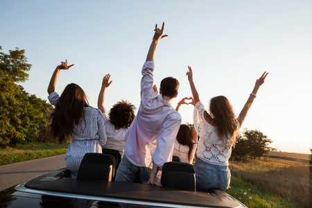Back view. Young guys are sitting and holding hands up in a black cabriolet on the country road on a sunny day.の写真素材