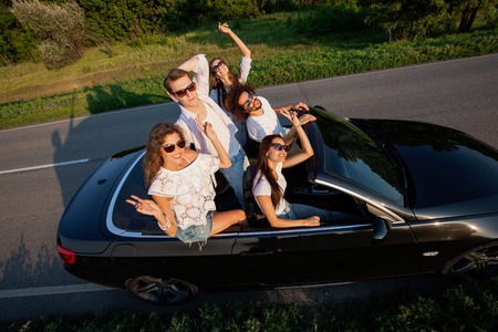 Stylish young guys are sitting and smiling in a black cabriolet on the country road on a sunny day.の写真素材