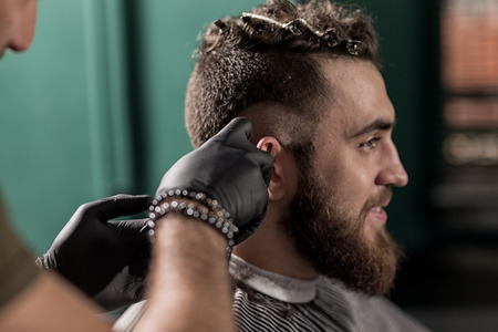 Brutal handsome man with beard sits at a barber shop. Barber shaves hairs at the side.