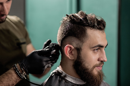 Brutal handsome man with beard sits at a barber shop. Barber shaves hairs at the side.の写真素材
