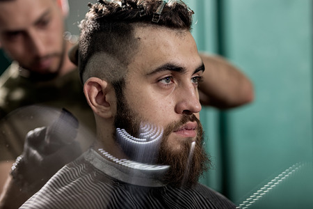 Handsome young man with beard sits at a barber shop. Barber shaves hairs at the back.の写真素材