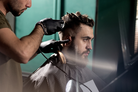 Handsome young man with beard sits at a barber shop. Barber in black gloves shaves hairs at the side.の写真素材