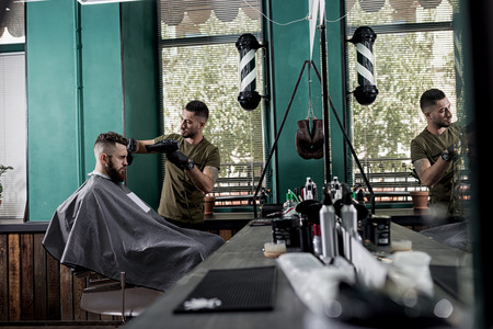 Young man with a beard sits in the chair in front the mirror at a barber shop. Barber dries mens...