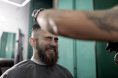 Stylish man with a beard sits and smiles at a barber shop. Barber in black gloves does the spraying for hairstyleの写真素材