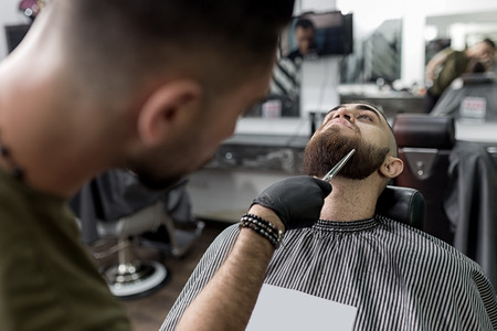 Stylish man with a beard sits at a barbershop. Barber trims mens beard with scissors.の写真素材