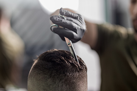 Barber in black gloves cuts with scissors hair on top of head of dark-haired man at a barbershopの写真素材
