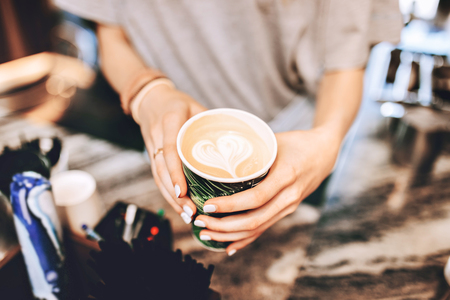 A youthful slim lady,dressed in casual clothes,is holding a coffee next to coffee machine in a lovely coffee shop.の写真素材