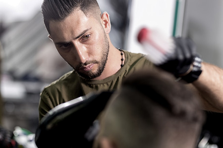 Handsome barber dries hair and makes a styling of dark-haired man at a barbershopの写真素材