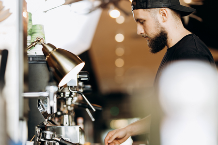 A stylish young man with beard,wearing casual clothes,cooks coffee in a coffee machine in a modern coffee shop.の写真素材