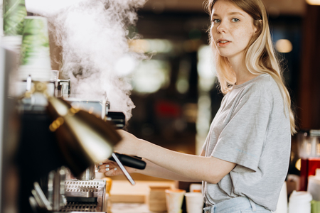 A youthful blonde slim gir,dressed in casual outfit,cleans the coffee machine with steam in a modern coffee shop.の写真素材
