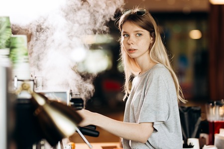 A youthful blonde slim gir,dressed in casual outfit,cleans the coffee machine with steam in a modern coffee shop.の写真素材