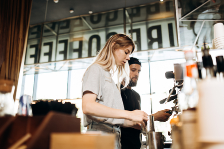 Two young baristas,a blonde girl and stylish man with beard,are shown cooking coffee together in a coffee machine in a cozy coffee shop.の写真素材
