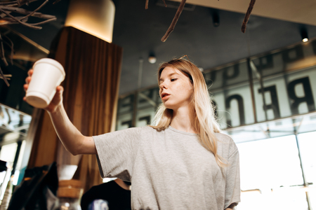 A youthful cute blonde girl,dressed in casual outfit,stands and holds a glass of coffee in a cozy coffee shop.の写真素材