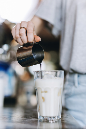 A youthful pretty slim blonde,wearing casual outfit, pours coffee into a glass of milk in a cozy coffee shop.の写真素材