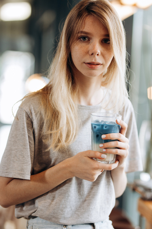 A good looking slim blonde  girl,wearing casual style, sits on the chair and holds a cup of coffee in a cozy coffee shop.の写真素材
