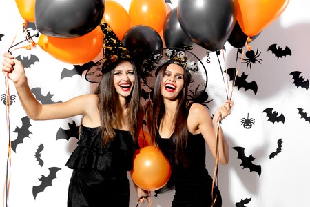 Two beautiful young women in witches hats are with black and orange balloons on a white background with black bats. Halloweenの写真素材