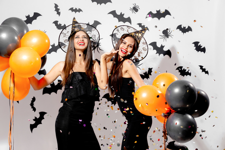 Two gorgeous women in witches hats smile and hold black and orange balloons on a Halloween background. Confetti aroundの写真素材