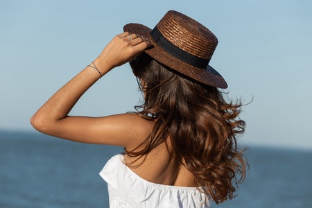 Portrait of gorgeous dark-haired girl  in white dress and hat near the sea on a sunny day. Back view.の写真素材