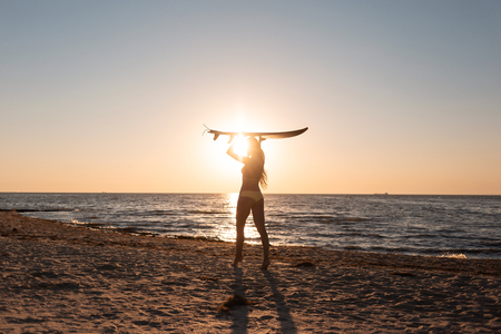 Girl in a swimsuit holds a surfboard over her head on the sandy beach near the sea on the sunsetの写真素材