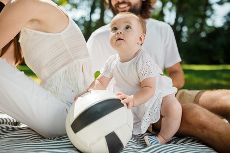 Adorable little girl in white dress sits next to parents on a blanket on the lawn and touches a...