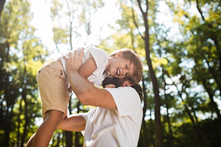 Dark-haired father with beard dressed in the white t-shirt is holding in the arms his blond son and smiling on a summer day in the park.の写真素材