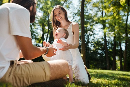 Charming little girl in a white dress is holding her mother by the hand and is trying to make her first steps towards her father on the green lawn.の写真素材