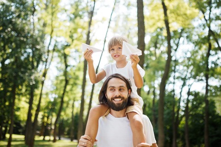 Father and his son walking in the forest. Boy dressed in a white t-shirt sitting on the shoulders of his father and keeps a paper airplane in his hand.の写真素材