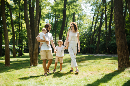 Warm sunny day. Handsome father, beautiful mother and two kids dressed in the white casual clothes happily walks in the park.の写真素材